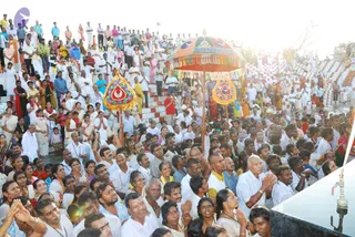 A large crowd of devotees fills red-and-white stepped ghats beside water, mostly dressed in white with some in colourful saris and salwar. Many stand with hands pressed together in anjali. Two ornate umbrellas with gold fringe and red-and-gold medallions rise above the crowd near the center, held aloft on poles. At the lower right, a metal fixture and tap sit on a dark platform edge.