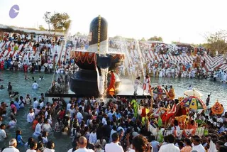A large black stone Shivalinga stands in shallow water at an outdoor ceremony, with water pouring over it from above and a garland of marigolds and red fabric draped around its base. Several people in white clothing and orange garments stand near the lingam in the water, and one figure in white with a red-and-white garment is visible beside it. Behind them, a dense crowd wades in the water and fills tiered seating, with many people in white. On the right bank, colourful umbrellas, a decorated cart with orange and red cloth, and people in bright saris and dhotis gather. The scene suggests an abhishekam in progress with a large crowd of devotees.