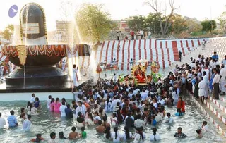 A large outdoor festival scene at a water-filled tank beside a tall black Shivalinga with orange and white horizontal bands and a central dark stripe, with water jets spraying from its base. Hundreds of devotees wade and bathe in the shallow water, many in white clothes, some in saffron, red and pink. A decorated ratha or palanquin with garlands and floral trim is carried on the right, surrounded by people. Red-and-white striped steps rise on the right, lined with spectators, and palm-like trees stand in the background under a pale sky.