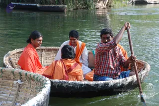 Outdoor photo of people riding a round woven bamboo coracle on a calm river. A man in a red-and-white checked shirt and dark trousers, standing at the stern, paddles with a long wooden oar. Four passengers sit in the boat: on the left a young man with hair tied back in a bun wears an orange robe; in the centre a person in orange robes faces away from the camera; a smiling man in a white shirt wears an orange shawl; another person behind wears orange. Reeds and rocks line the riverbank. a person's presence is not clearly established from appearance, so the a person field is unsure.