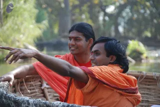 Outdoor photo of two men seated in a woven bamboo-and-wicker coracle boat on a river, surrounded by green foliage and trees. The man in front, wearing an orange saffron robe with a maroon shawl, extends his right arm to point toward the left while smiling. Behind him, a second man in a red-orange robe looks on with a smile. The boat has a woven basket-style hull with rope lashings.