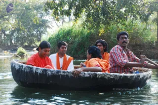 A photograph of people riding in a dark round wicker-and-pitch coracle boat on a river with green riverbank vegetation and trees behind. Four people are visible: a woman on the left in an orange-red garment smiling, a man in a white shirt with an orange shawl seated beside her, a young man in an orange shawl at the back, and a man in a red-and-white checked shirt rowing with a paddle on the right. A second woman in orange clothing sits in the middle of the boat, trailing a hand in the water. The water is calm and green-brown.