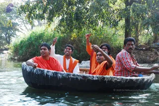A photograph of five men riding in a dark wooden coracle boat on a calm river, with green riverbank foliage and trees behind them. Four men wear saffron-orange garments; the man in the centre-left wears orange with a red wrap, a man behind him wears white with an orange shawl, and the man on the right wears a red-and-white checked shirt. Several raise their arms and point upward, appearing to chant or call out. No identifiable a person candidate can be confirmed from appearance, so roles are described as devotees and passengers in the boat.