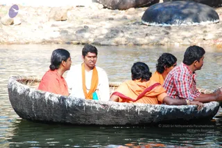 Five people ride in a dark dugout coracle boat on calm water, with large dark rounded rocks and a rocky shore behind them. A woman on the left wears an orange garment and has hair pulled back. A man in white clothing with an orange shawl sits in the centre-left looking to his side. Another person in an orange garment with a maroon edge sits with their back to the camera, and two more men sit at the right, one in a red checked shirt. The boat is white-streaked and bundled with rattan. No identity of any individual is asserted, and no candidate with distinctive a person cues is clearly present.