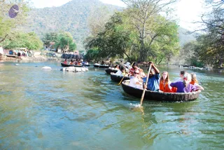 A photograph of several round wooden coracle boats moving in a line along a green river. Passengers sit in the boats in orange, white and purple clothing, and a man in a blue shirt stands in the foreground boat paddling with a long wooden oar. Forested hills and tall trees line the riverbank, and a small group of onlookers stands on the far shore near buildings. Several people are visible in the boats, but no one can be identified as a person with confidence.