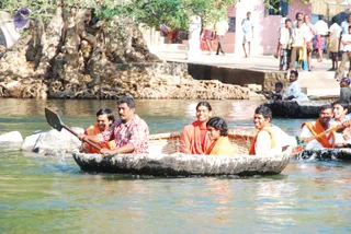 A photograph of a round wooden coracle boat on a river at Hogenakkal, carrying several people. In the boat, a man in a red-and-white checked shirt paddles, a man in an orange life vest sits at the left, and a person in orange robes sits centre-right beside a young person in orange. A man in a white shirt and orange life vest sits at the right. Other people in orange vests are in a second boat at the right edge. Behind them, a rocky bank with exposed tree roots and spectators in dhotis, saris and shirts stand on a concrete ghat and steps near a pink building. The people in the boat are on the water, not on a stage.
