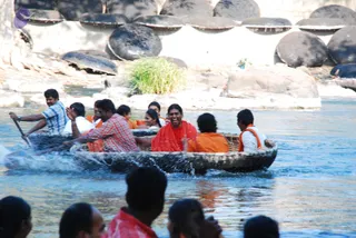 Outdoor photo of a group riding a round wicker-and-wood coracle-style boat on a river, with water splashing around the hull. A woman with long dark hair in a red-orange shawl laughs broadly at the centre of the boat, while a man in a red checked shirt and a man in a light blue shirt paddle. Other passengers wear orange and white garments and orange life vests, and a man in an orange vest sits at the right edge. Large dark rounded boulders and a grassy bank form the background, and several onlookers in the foreground watch from the shore.