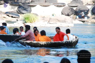 A photograph of people riding a round wooden coracle boat on a river, with rocky shoals and rounded stone formations along the far bank. Several men and women in saffron and orange clothing sit in the boat, some in white shirts with orange sashes, smiling and talking. A man in a red-and-white checked shirt at the left back of the boat steers with a paddle, while another man in a red shawl sits center right. Behind them, others are visible on the bank and in another boat, and dark silhouettes of spectators line the bottom edge of the frame.