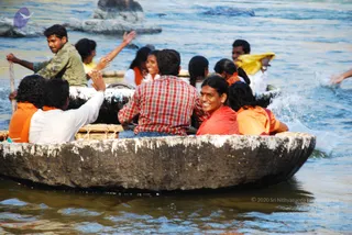 A photograph of several people riding in a large round wooden coracle boat on choppy, sunlit river water. Passengers wear orange, saffron and white clothing; one smiling young man in a red shirt sits at the centre facing the camera, while a man in a checked red shirt sits beside him. Another man in olive-green stands at the left holding a long oar, and others raise their hands and wave. Water splashes at the right edge. The setting appears to be a river with rocks in the background, consistent with a boat outing.