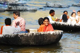 Outdoor photo on a river with rocky outcrops and brownish water. Several people ride round black coracle-style boats, woven-rim wooden bowls. In the foreground, a smiling man with short dark hair wears an orange-red robe and sits in a coracle, looking toward the camera. Beside him, a person in an orange garment sits low in the boat. A man in a white shirt and a man in a red-and-white checked shirt sit in the rear boat, and women in white and orange clothes sit in the other boat behind.
