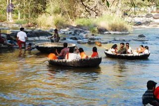 A photograph of round wooden coracle boats on a river with rocky banks and dry grass. In the foreground boat, several people in orange and white clothing sit, including a person in an orange top and white shirt; in the right boat, passengers in orange, white and red garments paddle with wooden oars while others sit at the front. A man in a red-checked shirt stands in the water beside the boats, another man in white shirt and red shorts wades nearby, and a man in a plaid lungi gestures from the rocks. Dense trees and boulders frame the scene under bright daylight.
