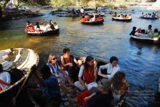 Several round woven bamboo coracle boats float on a brown river among rocks, each carrying a group of passengers. In the foreground boat, a man in a blue shirt and green lungi paddles while women in orange and white sit beside him. Other passengers wear white kurtas, orange scarves and saffron shawls. A Western woman in sunglasses and a light blue top sits in the front. The bank and dry trees are visible at the top, with other boats and people in the background.