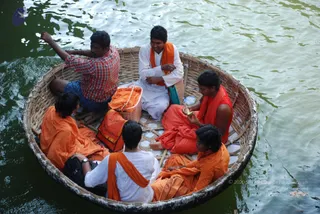 Photograph of a round woven bamboo coracle boat floating on dark green river water at Hogenakkal, with six people seated inside. Several wear saffron-orange robes and orange shawls, while two men wear white kurta-style shirts with orange shawls, and one man in a red-and-white checked shirt sits at the back raising his arm. A woman in orange draped clothing peels a fruit, and an orange bag and white plastic basket sit among the group.