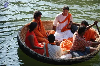 A photograph of a round woven bamboo coracle boat on greenish river water. Six people sit inside: a woman in a red garment with a red shawl at left, a young man in white kurta with an orange shawl standing at the top centre, a man in orange sitting beside him, a man in a red checked shirt and jeans rowing at right, and another rower in orange at the back right. A child-sized figure in orange sits near the front, and a wicker basket holds orange cloth. a person's appearance cannot be confirmed here, so no candidate is identified.