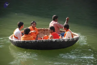 A photograph of a round wicker coracle boat on green river water at Hogenakkal Falls. Six people sit inside. Most wear orange-saffron shawls or wraps over white or orange clothing. A man in a red-and-white checked shirt at the back of the boat raises one arm and steers, while a young man in a white kurta with an orange shawl sits at the front right. A man in an orange shawl at centre left smiles. A woman in a red top sits in the middle. Water ripples around the dark rim of the boat. No identity of any person is asserted.