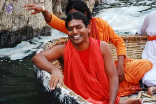 A photograph of two men riding a woven bamboo coracle on rippling river water beside rocky cliffs at Hogenakkal. In the foreground a smiling man with shoulder-length dark hair and a red tilak wears a red shawl over a red top and rests his arm on the boat's rim. Behind him a man in orange robes with a red sash raises one arm, and at right a person in white clothing sits on the boat's edge.