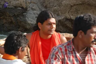 Outdoor photograph on a rocky riverbank beside water. A man with long dark hair tied back, a vertical tilak on his forehead, a red-orange shawl over a red sleeveless top sits in a wooden boat or raft. Two other men are partly visible in the foreground: a young person in a white garment with an orange cloth, and a man in a red and white plaid shirt with a moustache. Swamiji (probable) is the man in the centre, identified by long swept-back hair, orange-saffron shawl and ochre clothing cues with a sannyasi-style tilak.
