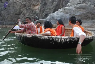 A photograph of a round wicker-and-pitch coracle boat on green river water, with steep grey rock walls behind it. A man in a red checked shirt stands at the stern rowing with a long wooden oar. Five people sit inside in orange and saffron garments: a young man in an orange wrap at left smiling, a person in orange wrap seated centrally with dark hair, a woman in a red-orange top at centre-right, and a young man in a white shirt with orange wrap at right, wearing a wristwatch. Another figure in orange sits at the far right edge. Another person stands on the rocks at the upper left. No clearly identifiable a person figure is present.