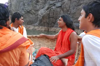 Photograph taken on a wicker-hulled coracle-style boat on water beside a tall rocky gorge wall. A young man with long dark hair, a bare-armed red sleeveless top and red-orange wrap sits centrally, one arm extended to the boat's woven rim, speaking with others. He has long swept-back hair and orange-red robes, cues consistent with the a person label. Around him sit three companions in orange and saffron garments and white shirts, one in left foreground with an orange wrap, another in right foreground in white with an orange shawl, and a man in white with an orange shawl at upper left. A black bag rests in the foreground and a pink plastic basket sits at left.