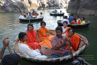 A photograph of devotees riding round wicker coracle boats on a river between tall rock walls. In the foreground boat, a man in a white shirt and orange shawl sits at left, a man in a red-checked shirt and blue lungi paddles at centre, and a man in an orange robe with an orange shawl paddles at right. Next to them, a young man in orange robes and a woman in red and orange sit in the boat, and a man in white holds a basket. Other boats behind carry more people in orange, white and yellow shawls. A small plastic basket and a camera are visible at the bottom.