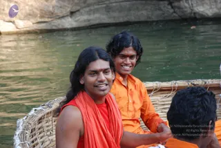 Outdoor photo on a river with rocky cliffs behind green water. In a woven bamboo basket boat, a smiling man with long dark hair, a red sleeveless top and a red-orange shawl wrapped over his shoulders sits at front-left, with a red tilak on his forehead. Beside him, a younger man with curly hair wears an orange shirt and smiles, his hand on the boat's edge. In the lower right foreground, another person with short dark hair is partly visible in orange. A purple logo appears in the upper left corner. Swamiji (probable) is the man with long hair in saffron-red shawl and orange-red clothing seated at front, shown by long swept-back hair, saffron shawl and ritual clothing, so the candidate is present in person.