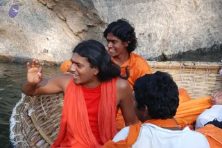 A photo of people in a round woven basket boat on calm water beside a large rock face. A man with long, swept-back black hair wears a red-orange shawl over a red top and gestures with his raised right hand while smiling. Behind him, a younger man with curly hair in an orange garment smiles. In the foreground, a man in a white top with an orange shawl sits with his back to the camera.