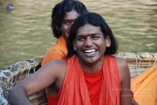 Close photo of a smiling man with long dark hair and a red tilak on his forehead, wearing an orange-red shawl over a red sleeveless top, seated in a woven bamboo boat on green river water. Behind him a second man in orange robes with a red mark on his forehead also smiles. a person: the first man has long swept-back hair and saffron-red sannyasi clothing, but the appearance is not conclusive enough for a firm identification, so the claim is recorded as an uncertain in-person candidate.
