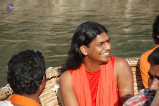 A smiling man with long black hair sits on a bamboo raft on a river, looking to his right. He wears a sleeveless red top with an orange-saffron shawl draped over his shoulders and a red mark on his forehead. Two other men are partly visible at the edges of the frame: one in the lower left with short dark hair, and one at the right in an orange shirt. Behind the raft is rippling green-brown water in bright sunlight.