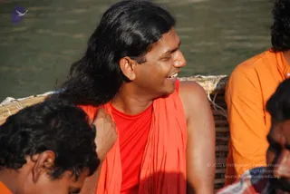 A smiling man with long, swept-back black hair sits outdoors in front of a green river, wearing a bright red sleeveless top under an orange-red shawl draped over his shoulders. Beside him, a man in an orange robe sits on a woven bamboo basket-like surface, and partly visible at the left edge is another person with curly dark hair. The man at centre wears a small mark on his forehead and looks off to the side, appearing cheerful. Appearance-based cues of long hair and a saffron-toned shawl suggest a person, though this is uncertain.