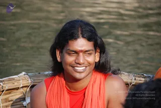A smiling man with long, swept-back dark hair and a red tilak on his forehead sits in front of a rippling river, wearing an orange-red shawl over a red sleeveless top. Behind him is a bamboo raft or platform lashed with white cord, and a blurred figure in the lower right corner. The shawl, long hair and tilak are cues for the Swamiji (probable) appearance-based label, shown here as a person seated on a bamboo raft during the Kodikkan Darisana yatra at Hogenakkal.