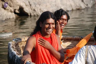 A photograph taken on water shows two men seated in a woven bamboo coracle-style boat, laughing broadly. The man in front, with long swept-back dark hair, wears an orange-red sleeveless garment wrapped over his shoulders and a red top, and has his hand resting on his chest. Behind him, a younger man with short wavy hair wears an orange cloth and cups his hand near his ear, also laughing. A paddle with an orange blade rests at the right edge beside a person in white. Pale rocky cliffs and rippling river water fill the background.
