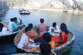 A photograph of several round wicker coracle boats on a river between steep pale rock cliffs. In the foreground boat, a young man in a white kurta with an orange shawl sits at the front beside a bearded man in an orange garment, while a woman in a red top and orange wrap sits at the rear; a man in a red checked shirt sits behind them and an oarsman in a green-and-blue checked shirt paddles the boat on the left. Other boats carry passengers wearing orange and white clothing, and a boatman in the distance steers. The scene shows devotees boating together on the water.