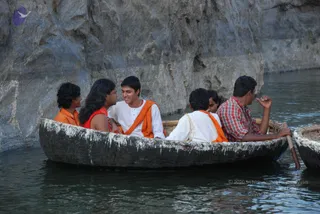 A photograph of a group riding a round wicker-and-pitch coracle boat on calm water beside a steep grey rock face. Six people sit in the boat: a young man in a white kurta with an orange shawl sits centre and smiles toward a long-haired person in a red top; a person in an orange top sits at left; a man in a white dhoti with an orange shawl sits near the stern; a man in a red-and-white checked shirt paddles at the right. The scene suggests a riverside boat excursion at a rocky gorge.