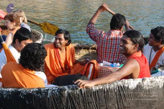 A photograph of a group of people laughing together on a coracle-style round boat with a black waterproofed rim, on a river at Hogenakkal. A man in the center wears an orange-saffron shawl and sits smiling beside a wicker basket with water bottles. Another man in a red sleeveless top grins in the foreground, and a young man in a white shirt with a rudraksha mala and saffron shawl sits at right. Behind them, a boatman in a red-checked shirt paddles with a long oar, and an older woman with reddish hair and a yellow paddle is seen at left. Water and rocks fill the background.