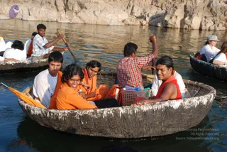 A photograph of several people riding in round wicker-and-pitch coracle boats on a river with a rocky cliff behind. In the foreground boat, a young man in an orange-saffron top and dark trousers sits near the front, next to a smiling man in orange robes who holds a small water bottle, while a man in a red sleeveless top sits at the right edge. A man in a white shirt sits at the left edge and a man in a checked red shirt paddles in the middle boat. A basket with a bottle and papers sits in the boat, and a woman in a grey cap and white shirt is visible in a boat at the right. Other people in white and orange clothing sit in nearby boats on dark water.