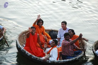 A photograph of a round woven bamboo coracle boat on open river water, with several smiling men crowded inside. A man in a loose orange-red shawl and orange clothing sits at the left front, waving with his right hand. Behind him a man in orange waves his hand high, and a young man in white kurta sits in the centre-back beside a dark bag. A man in a red-and-white checked shirt and blue jeans paddles at the front right, and another man in orange stands at the right edge. Two more paddles and a boat are partly visible at the left and right edges. A small purple bird logo appears in the upper left corner.