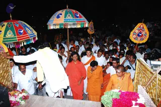 Night procession in a crowded Indian street. Several umbrellas with bright multicolored panels and blue dove-like emblems, plus the word Dhyanapeetham, shade the scene. A man in a white shirt and cream shawl bends forward with an arm outstretched toward a golden platform. In front, a man in a rust-red robe and a second man in orange cloth walk beside a woman in orange who covers her face with her hand. A bald man in orange wears a beaded necklace. Beds of flower petals in pink and green lie at the platform's base, and a large crowd in white and orange surrounds them.
