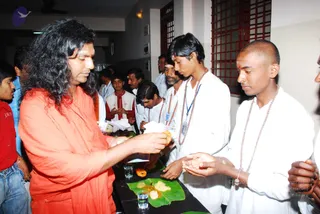 Indoor photo of a man with long, swept-back wavy black hair and a red-orange wrap, standing at a table and serving food from a banana leaf to a younger devotee. Several devotees in white clothes and rudraksha malas stand around the table, one holding out his hands, another in a white shirt with a blue lanyard. A small glass of water and food items such as fried snacks and rice sit on the leaf. Other people stand in the background near a red-grilled window.