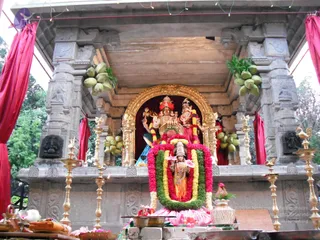 A photograph of a decorated shrine structure with stone pillars draped in red cloth and hung with green coconut clusters. Inside a gilded arch stands a deity murti, a seated figure in red crown and ornate attire beside a standing consort in pink, with a small standing figure in orange garments decked in marigold and green leaf garlands placed on a pink-draped pedestal. Brass oil lamps stand on the ledge, with brass vessels, flowers, and offerings in the foreground. No people are visible.