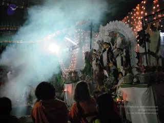 Night photograph of a Navaratri puja at an illuminated shrine. Smoke or incense haze drifts across the frame in front of a decorated deity murti of Durga with multiple arms and a white lace-style backdrop, flanked by smaller figures and surrounded by fairy lights. Several people seated from behind in orange-brown clothing face the shrine, with a white-clad figure working near the left. Flower arrangements and oil lamps sit on the altar tables, and a banner with lettering is visible at the top right.