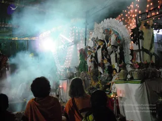 Night-time photograph of a Durga-style deity murti with a white lace-like arch and tiered white decorations, flanked by smaller figures and surrounded by fairy lights and potted plants. Smoke or incense haze fills the left side, lit by a bright lamp. In the foreground, three people seen from behind sit facing the shrine: two wear orange-saffron wraps and one wears a dark top. A small brass lamp stand with a lit flame stands at the right near the altar, and a figure in white is partly visible in the haze at the left.