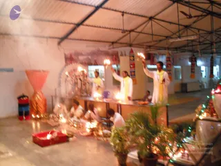 A blurred night-time photograph of an indoor aarti in a covered hall with a corrugated roof and ceiling fans. Three standing men in white dhotis and yellow shawls raise lit oil lamps toward a decorated altar with a deity murti. Seated devotees in white sit on the floor at lower left, near glowing oil lamps, a red tray, and potted plants. Strings of coloured lights decorate the right edge, and hanging banners with patterns stand along the wall.