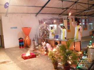 A photograph of an indoor puja in a corrugated-roof hall. Three standing priests in white kurtas with yellow stoles raise lit oil lamps toward a decorated deity murti on a white altar, a goddess figure with a crown and multiple arms flanked by smaller figures. Two or three seated devotees in white and pink sit at a low table holding brass plates and lamps. A red metal fire pit with a burning flame sits on the floor in front, a tall orange cloth-wrapped pillar stands at left, and potted plants and string lights decorate the right. Banners hang on the back wall.