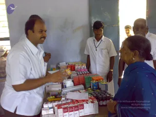 A photograph inside a plain room with pale blue-grey walls and a grey-green door frame. A man in a white shirt and trousers stands at left, gesturing toward a table covered with medicine boxes, bottles, tins and blister packs. Opposite him an older woman in a blue sari with a dark bun sits at right, listening. Behind the table a younger man in a white shirt with a lanyard stands, and two more men in white shirts are partly visible at the right edge near the doorway.