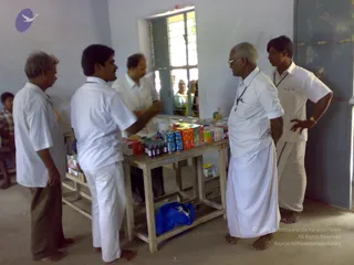 A photograph inside a bare, whitewashed room with a grey metal door and a barred window. Five men in plain white shirts and white dhotis stand around a rough wooden table stocked with medicine boxes, blister packs and small brown bottles. A man with grey hair and glasses, wearing a lanyard, stands at right with hands behind his back, while a younger man at right rests a hand on his hip. Two men in the window area and a child at far left look on. A blue plastic bag sits under the table. No one matching a person candidate is visibly identifiable from appearance, so the figures are described by role and clothing only.