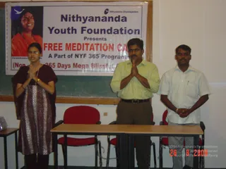 Three people stand in front of a poster board at a Nithyananda Youth Foundation free meditation camp. A woman on the left in a maroon shawl and patterned tunic with glasses and bangles stands with palms pressed together, a man in the centre in a yellow-green shirt and brown trousers with a belt stands with palms joined, and a man on the right in a white shirt and grey trousers stands with hands clasped. Two red chairs and a table sit in the foreground. The poster reads Nithyananda Youth Foundation Presents Free Meditation Camp, A Part of NYF 365 Programme, and beside it is a portrait labelled Paramahamsa Nithyananda, showing a smiling figure in orange clothing with a bead necklace.