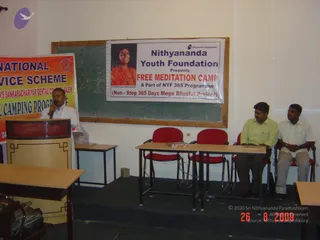 A small classroom-style hall with a man in a white shirt speaking into a microphone at a wooden lectern on the left, beside a large orange-and-yellow banner reading part of "...NATIONAL...VICE SCHEME" and "...CAMPING PROGRAMME". Behind him is a green chalkboard with a white banner reading "Nithyananda Youth Foundation Presents FREE MEDITATION CAMP A Part of NYF 365 Programme" with a portrait labelled "Paramahamsa Nithyananda" showing a smiling woman in orange clothing with a beaded necklace. Two seated men in a light green shirt and a white shirt sit on a low stage at right, with red-cushioned chairs and a small table holding a dish. Red date stamp reads 26 8 2009.