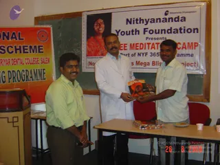 Three men stand in a classroom-style room before a banner reading Nithyananda Youth Foundation Presents Free Meditation Camp. A portrait labelled Paramahamsa Nithyananda shows a smiling woman in orange clothes. At left, a man in a light green shirt and dark trousers stands beside a lectern. In the centre, a man in a white lab coat holds a booklet with an orange cover and another man in a white shirt holds the other end of it, while both look at it. A table in front holds a box and a plate of food. A red-and-orange banner for a dental college programme hangs at left, and a chalkboard is behind them.