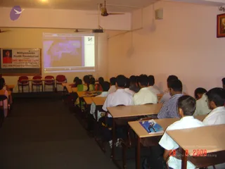 A classroom-style hall with rows of wooden desks and red chairs where students sit facing a projector screen showing a dim, grainy image. Most attendees are young men in white shirts and dark trousers seen from behind, with a few women in colourful salwar kameez at the left. On the left wall a banner reads 'Nithyananda Youth Foundation Presents FREE MEDITATION CAMP, A Part of NYF 365 Programme (Non-Stop 365 Days Mega Mission Project)' with a small portrait of a man in orange and red clothing. A framed picture hangs on the right wall and a ceiling fan turns above. The room is lit by overhead lights and a camera timestamp reads 26 8 2009.