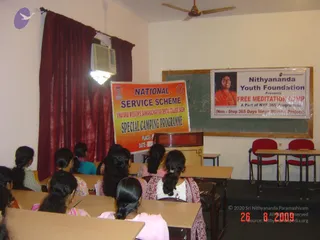 A classroom with wooden desks and red-seated chairs at the front, where a group of young women sit facing the front with their long dark hair in braids. Several wear salwar kameez in pastel and pink tones, and one wears a red and white patterned top. A red curtain hangs on the left and a ceiling fan turns overhead. A banner reading NATIONAL SERVICE SCHEME, VINAYAKA MISSION'S SANKARACARIYAR DENTAL COLLEGE, SALEM, SPECIAL CAMPING PROGRAMME hangs in front of a green chalkboard. Beside it, a poster shows a smiling woman in orange and reads Nithyananda Youth Foundation presents FREE MEDITATION CAMP. A wall-mounted air conditioner sits in the upper left.